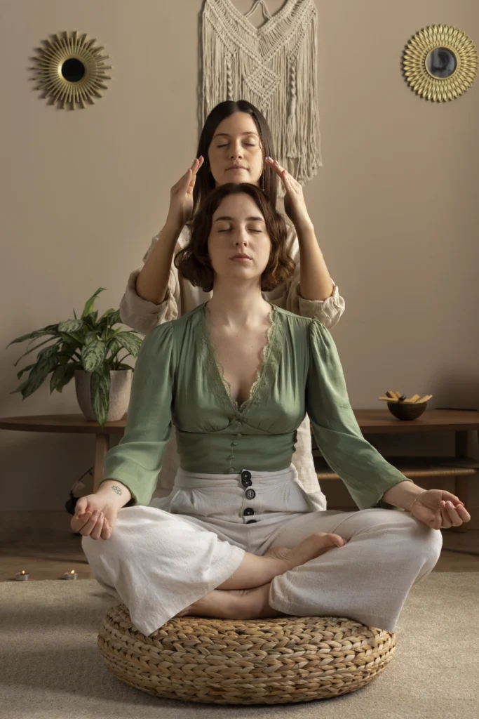 Une femme en position de médiation en premier plan, derrière elle une thérapeute canalisant l'énergie avec ses mains autour du visage de la patiente.